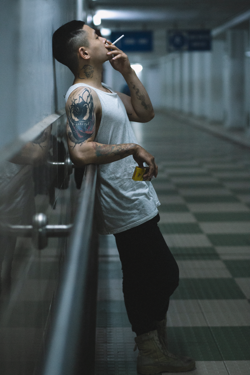 Free Side view of a tattooed man leaning against a wall, smoking indoors with a moody ambiance. Stock Photo