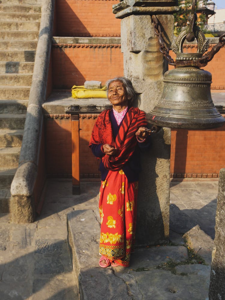 Old Woman With Red Shawl Standing Near A Bell