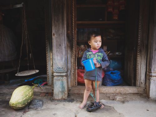 Free Child standing barefoot in a doorway holding a blue bucket, surrounded by rustic decor. Stock Photo