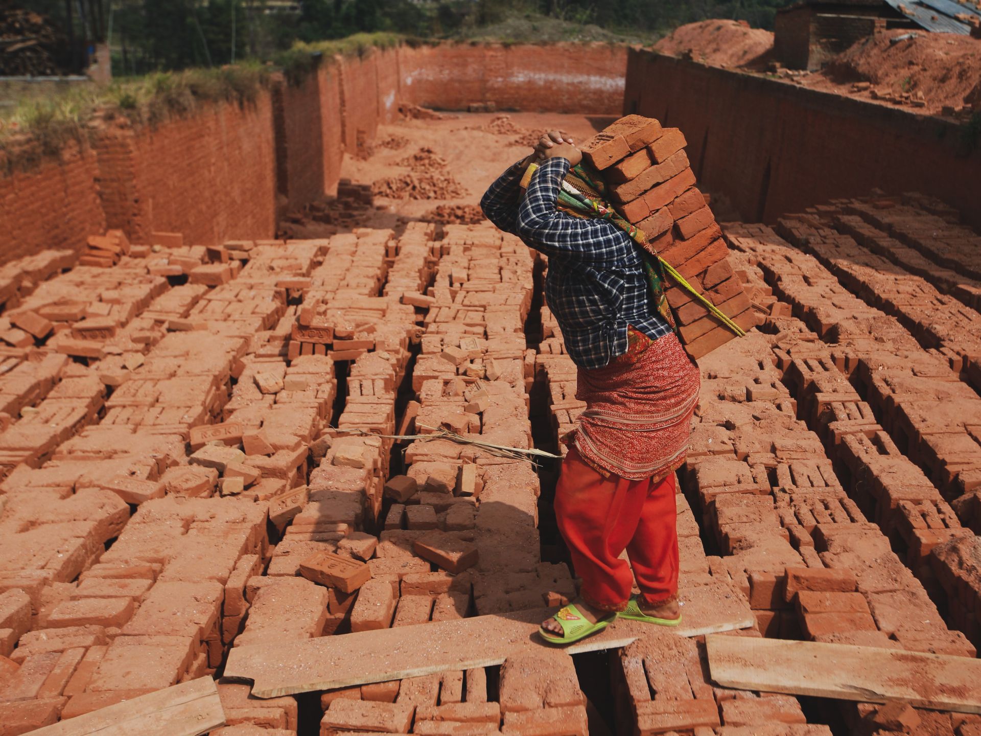 Woman working in a brick factory carrying heavy stacks of bricks outdoors, showcasing labor and industry.