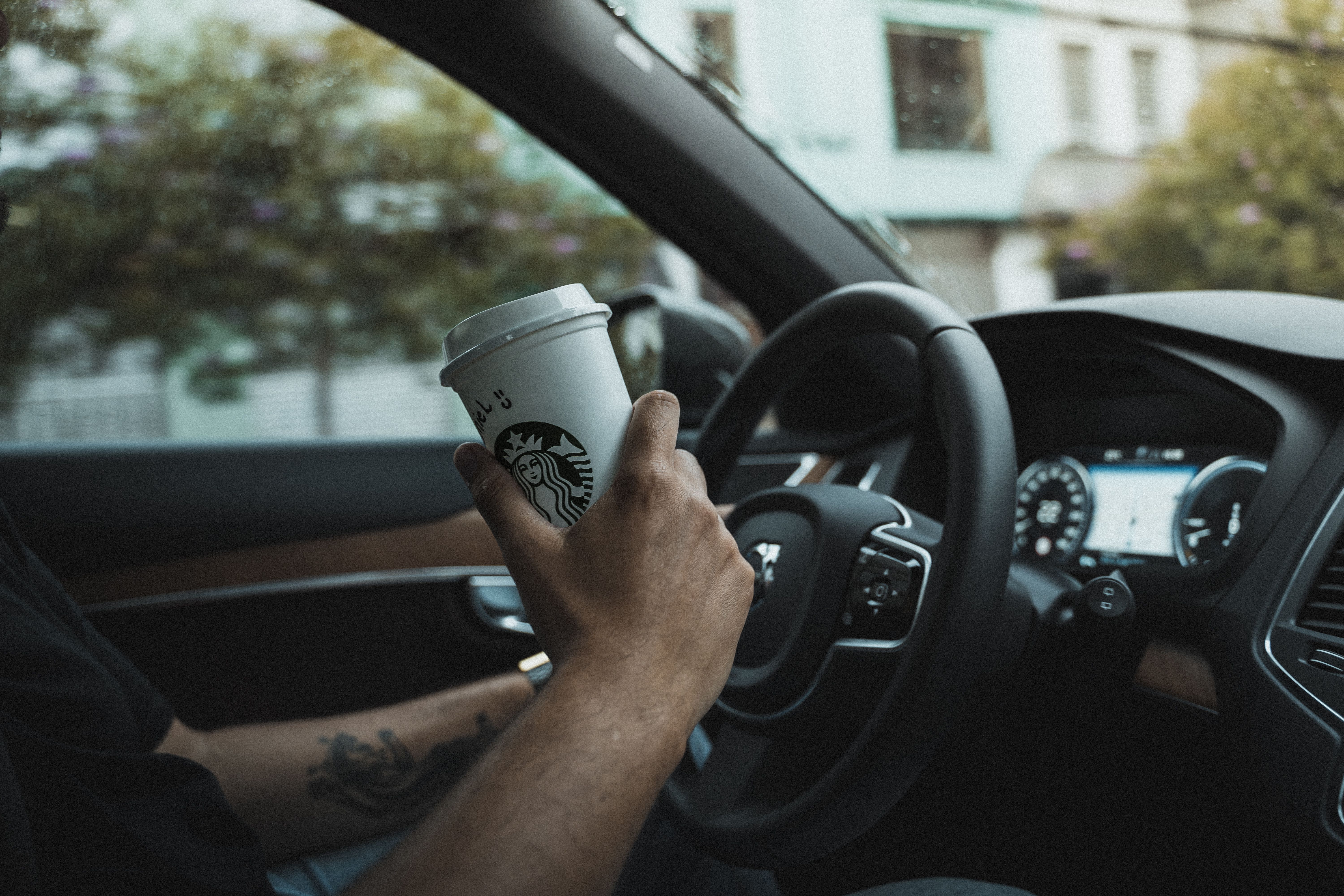 Tattooed hand holding a coffee cup inside a car, displaying urban driving lifestyle.