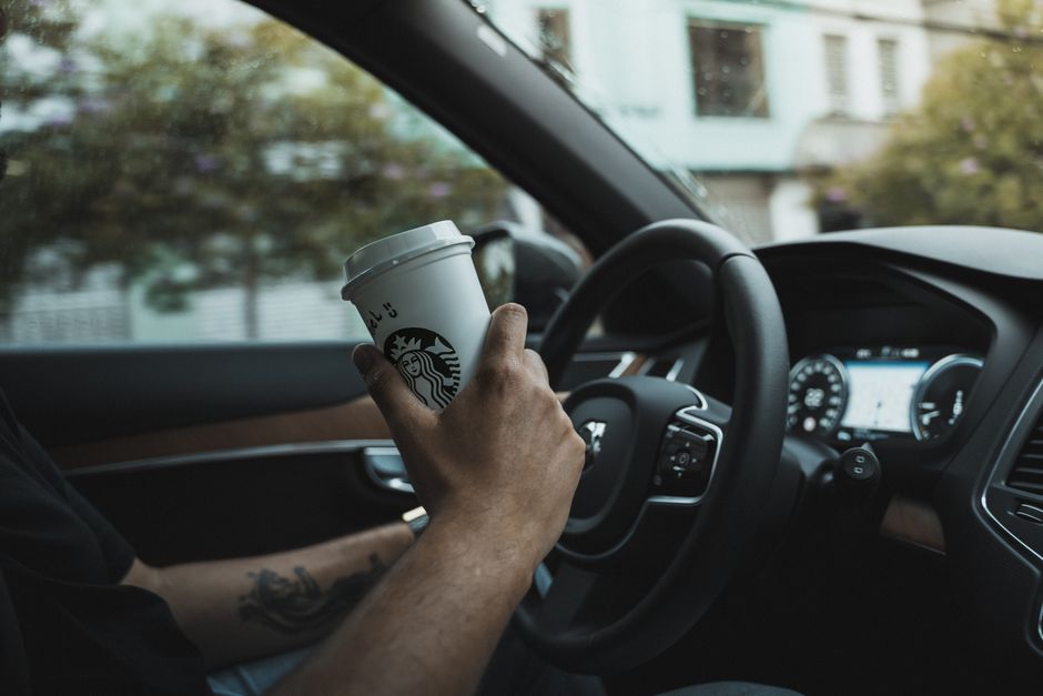 Tattooed hand holding a coffee cup inside a car, displaying urban driving lifestyle.