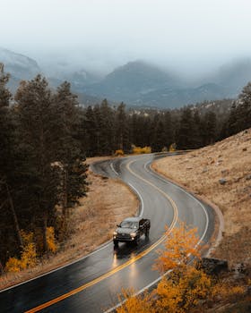 Car driving on a winding mountain road in autumn, surrounded by fog and vibrant foliage.