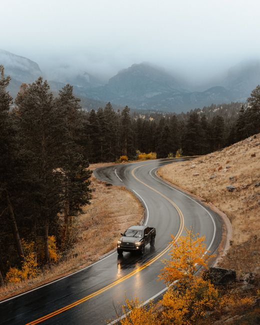 Car driving on a winding mountain road in autumn, surrounded by fog and vibrant foliage.
