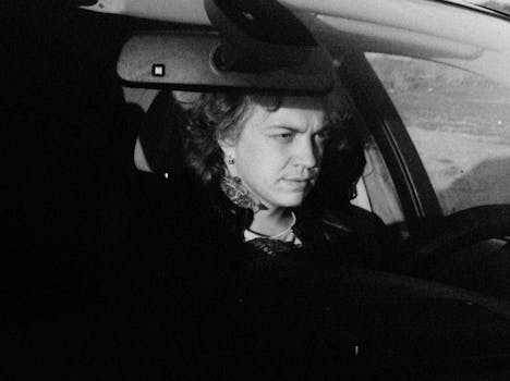 A black and white photo of a tattooed woman driving a car, capturing her in a thoughtful moment.