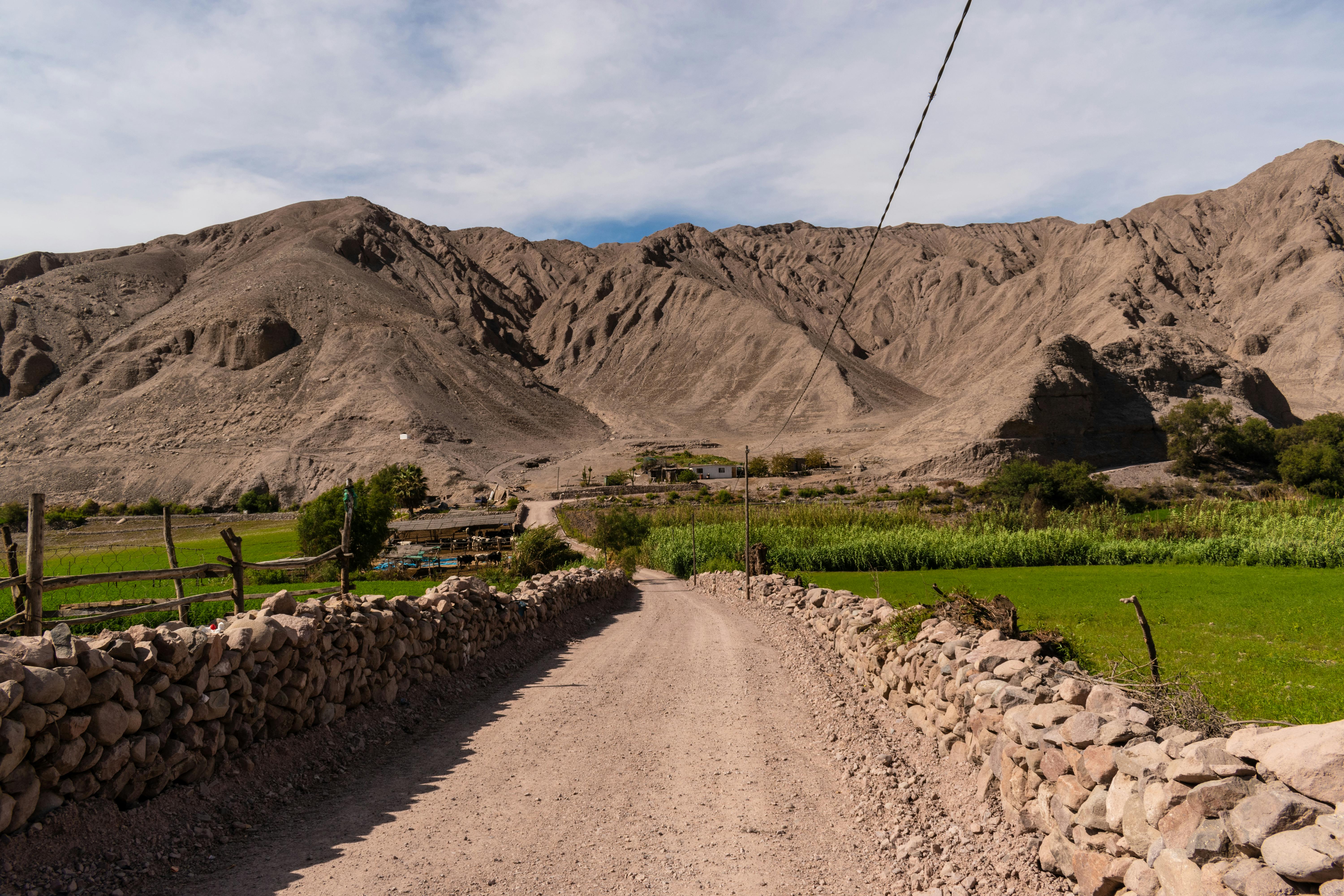 Picturesque dirt road leading through lush farmland and rugged mountains.