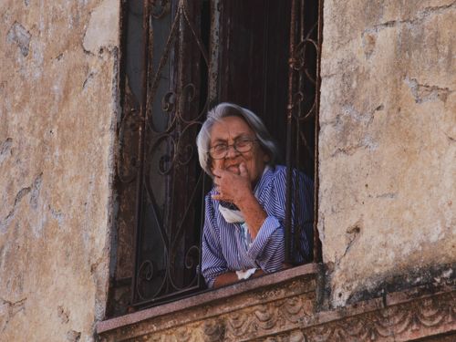 Free A senior woman looks pensively through an aged window with iron bars, embodying contemplation and solitude. Stock Photo