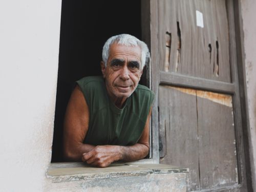 Free Elderly man in green top smiling and leaning out of a window with a wooden frame. Stock Photo