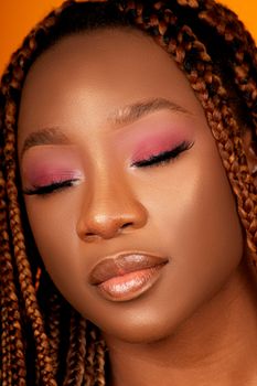 Close-up portrait of a woman with dreadlocks and vibrant makeup against an orange background.