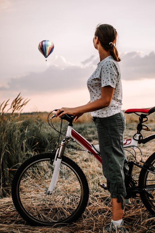 Free Woman standing with a bike in a field, watching a hot air balloon at sunset. Stock Photo
