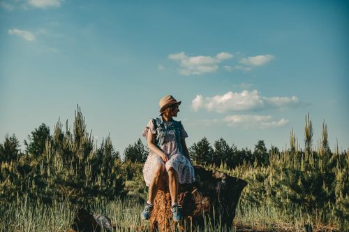 Free A woman in a floral dress and hat sits on a tree stump in a sunlit meadow during summer. Stock Photo