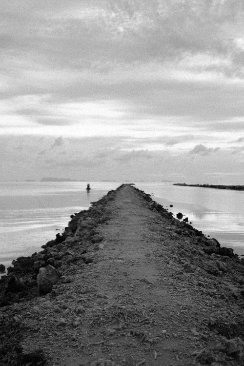 Free Black and white photo of a rocky path stretching into the calm sea under a cloudy sky. Stock Photo