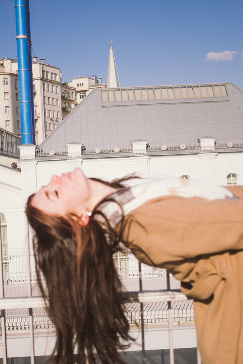 Free A woman in a brown coat leans back outdoors with urban buildings in the background, under a clear blue sky. Stock Photo