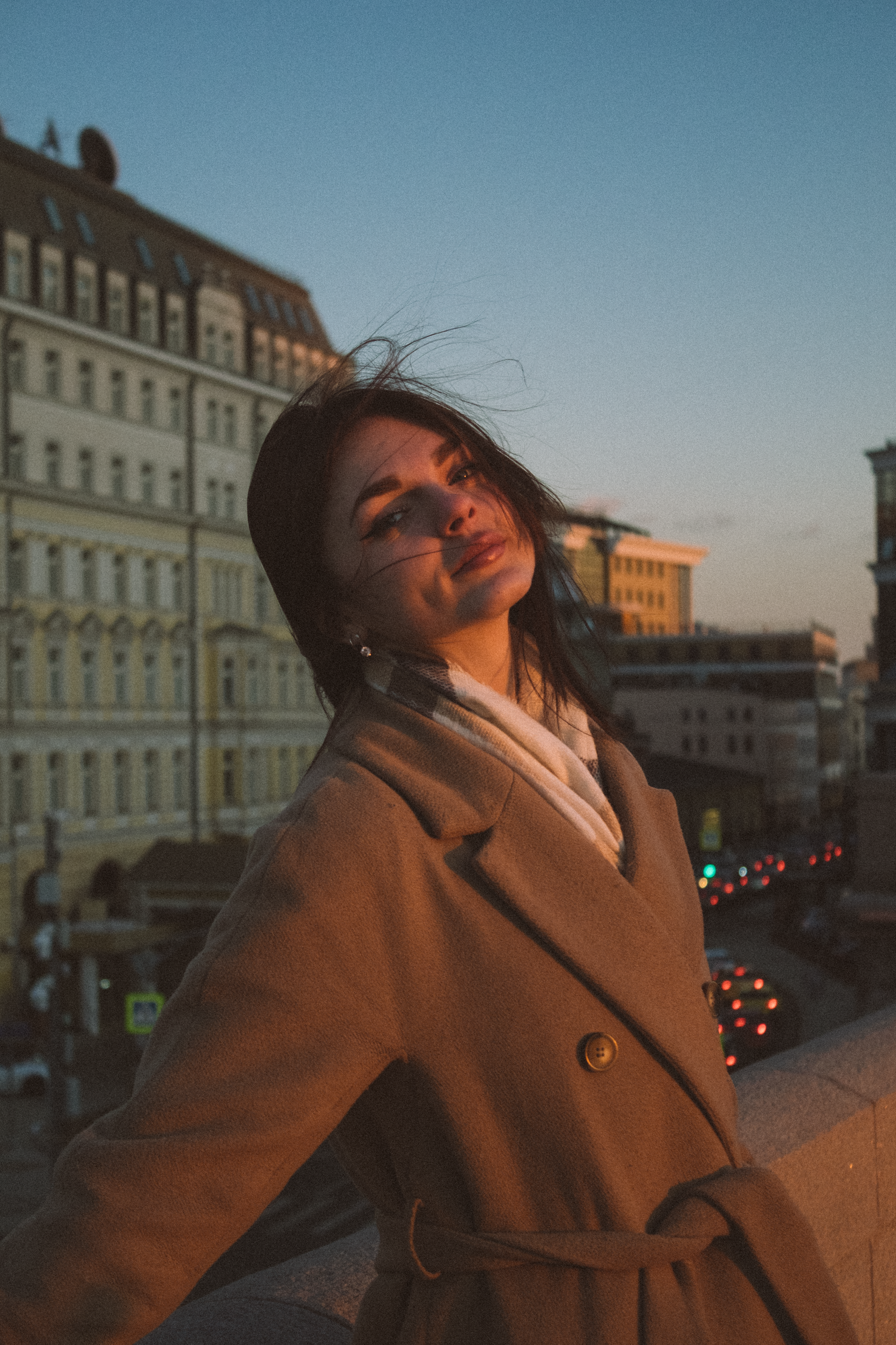 Woman in coat poses against city backdrop at sunset, exuding confidence.