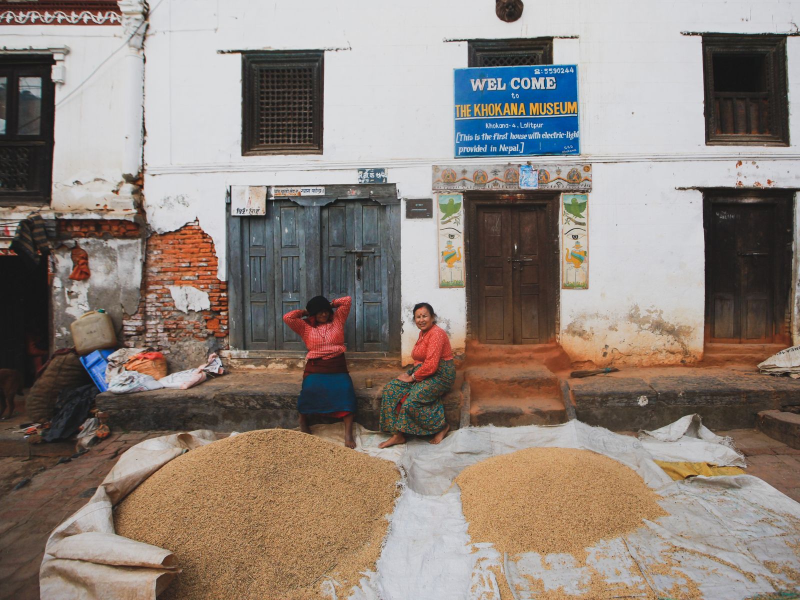 Grain drying in piles on sheets in the square at Khokana, two women resting on the step of a whitewashed house