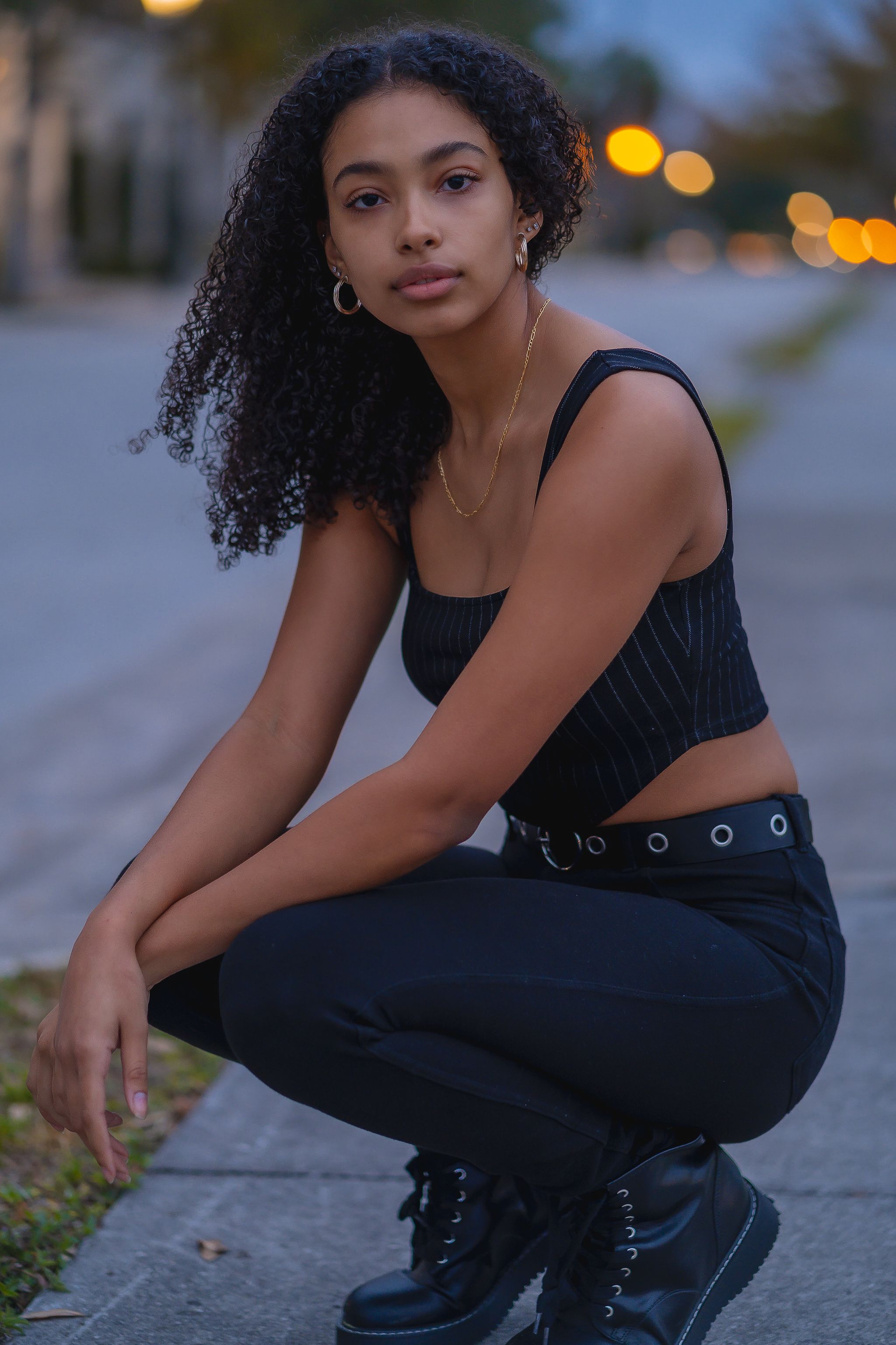 A fashionable young woman crouches on a sidewalk in a stylish outfit, showcasing modern street fashion at dusk.