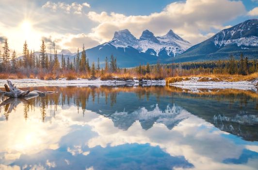 Stunning view of Three Sisters peaks reflecting on placid river at sunrise in Canmore, Alberta.