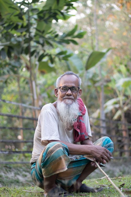 Free A senior man with a beard sits outdoors in a rural area of Bangladesh, exuding wisdom. Stock Photo