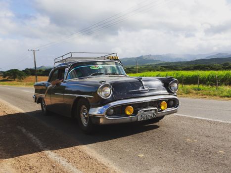 Classic vintage taxi car driving along a scenic rural road under a bright sky.