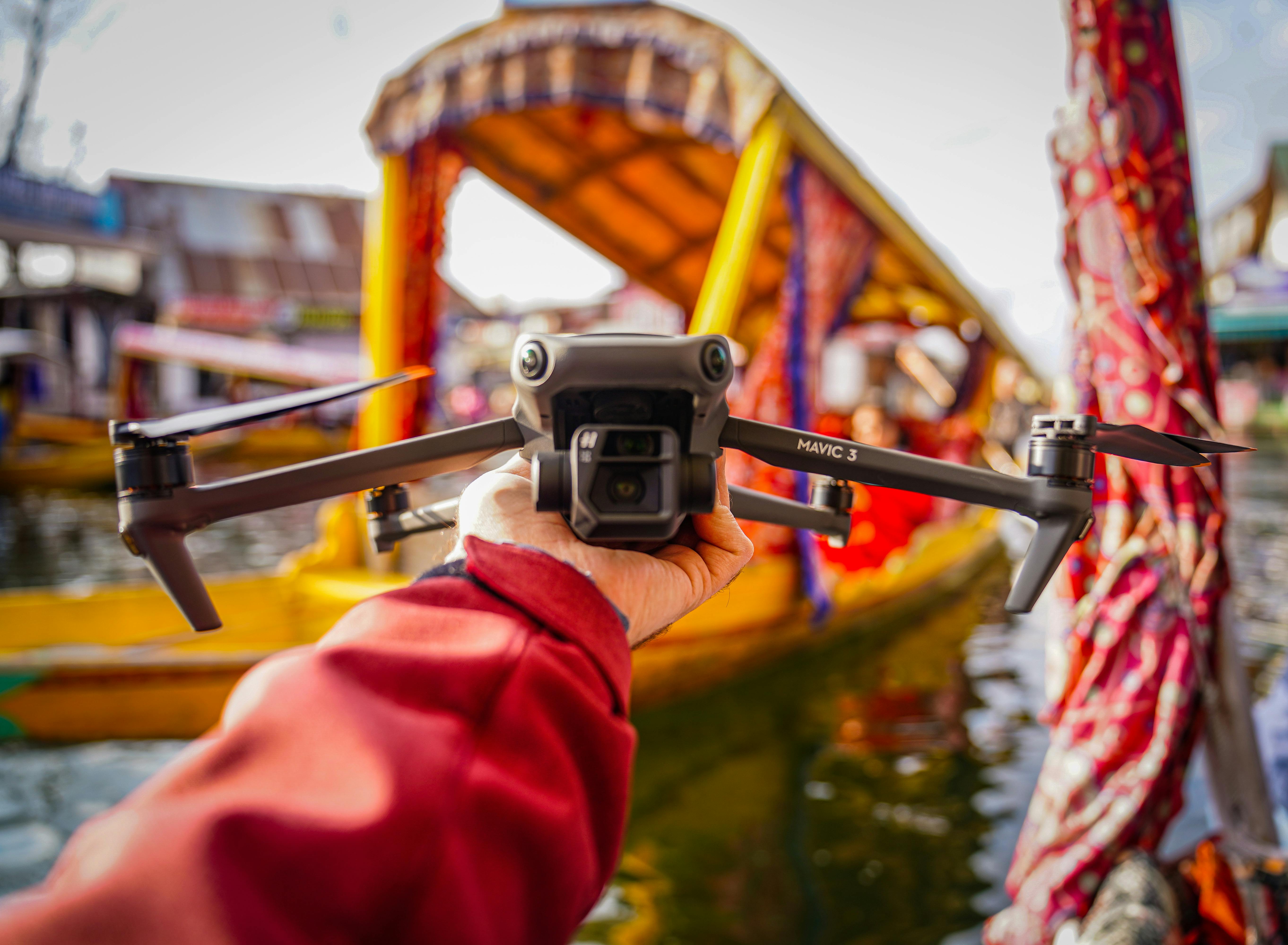 A hand holds a drone against a vibrant backdrop of colorful boats, showcasing travel and technology.