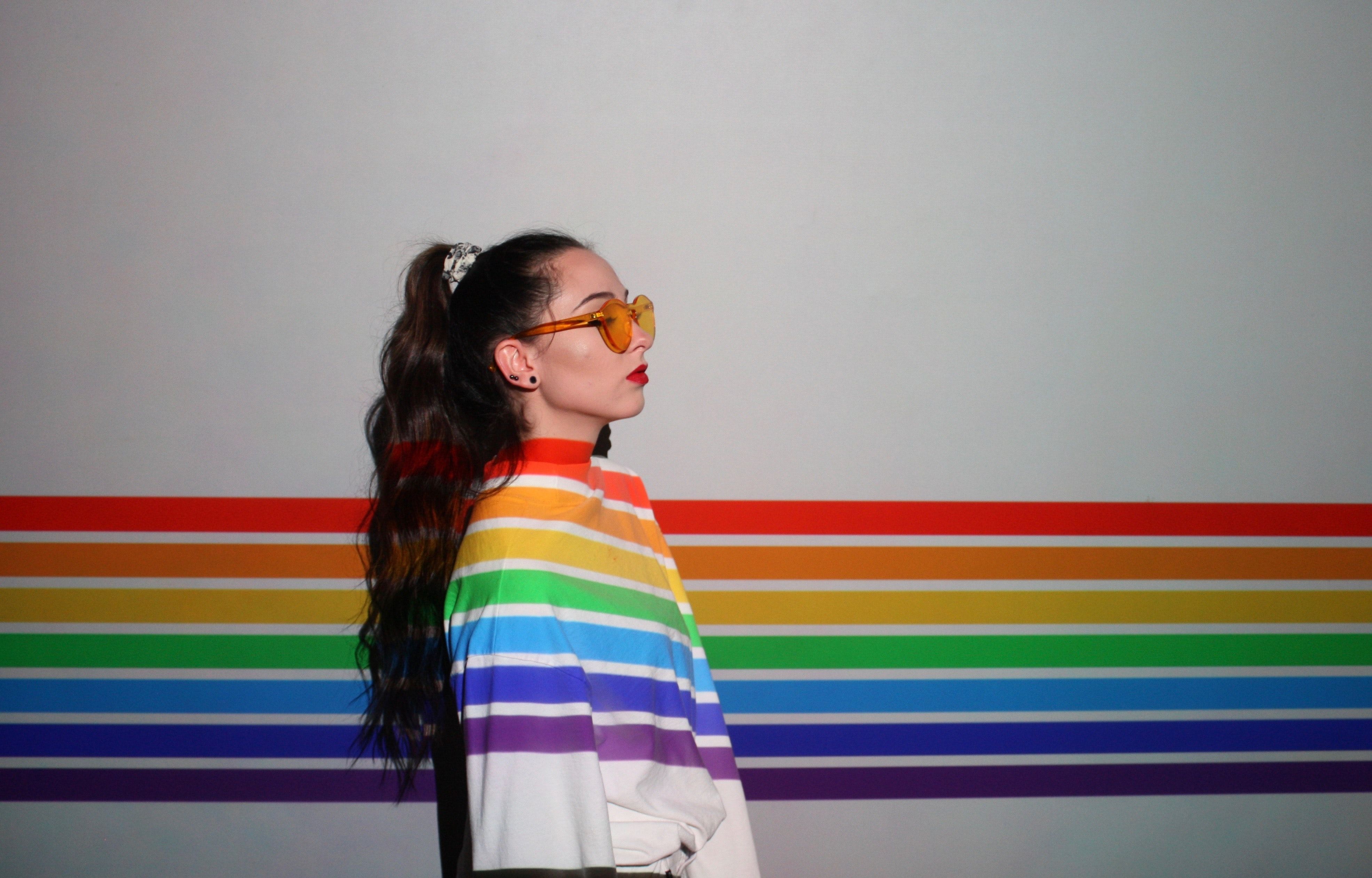 Side profile of a young woman in rainbow attire standing against a colorful striped wall.
