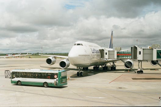 A large airplane docked at an airport gate with a shuttle bus nearby under cloudy skies.