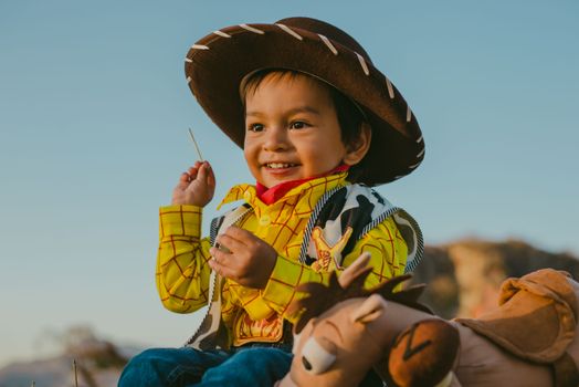 A cheerful child dressed as a cowboy enjoying an outdoor moment with a toy horse.
