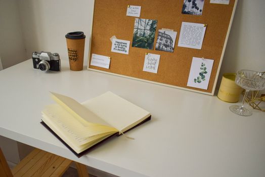 Cozy and inspiring workspace featuring a cork board, open notebook, and vintage camera on a white desk.