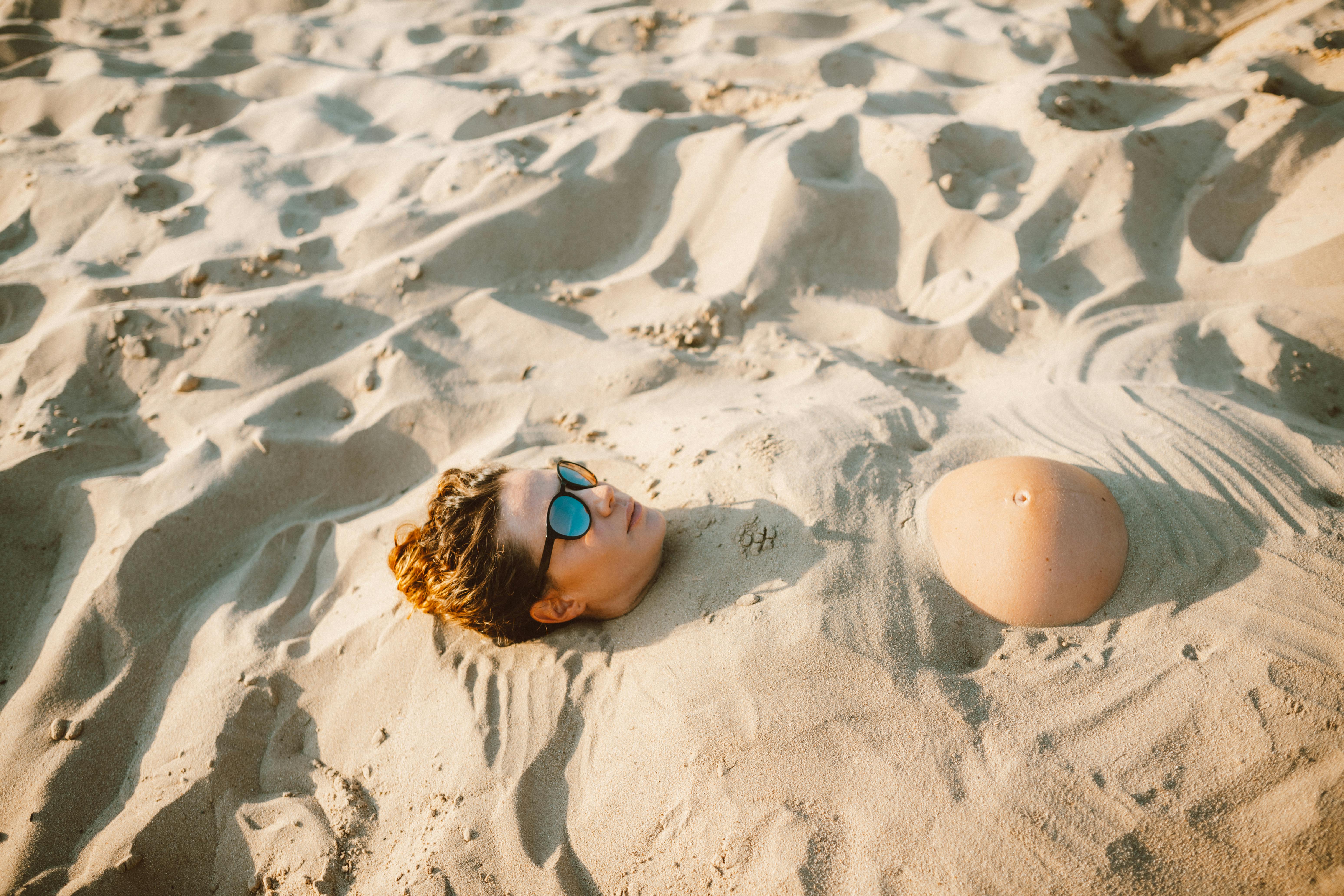 Person wearing sunglasses buried in white sand, enjoying a sunny beach day.