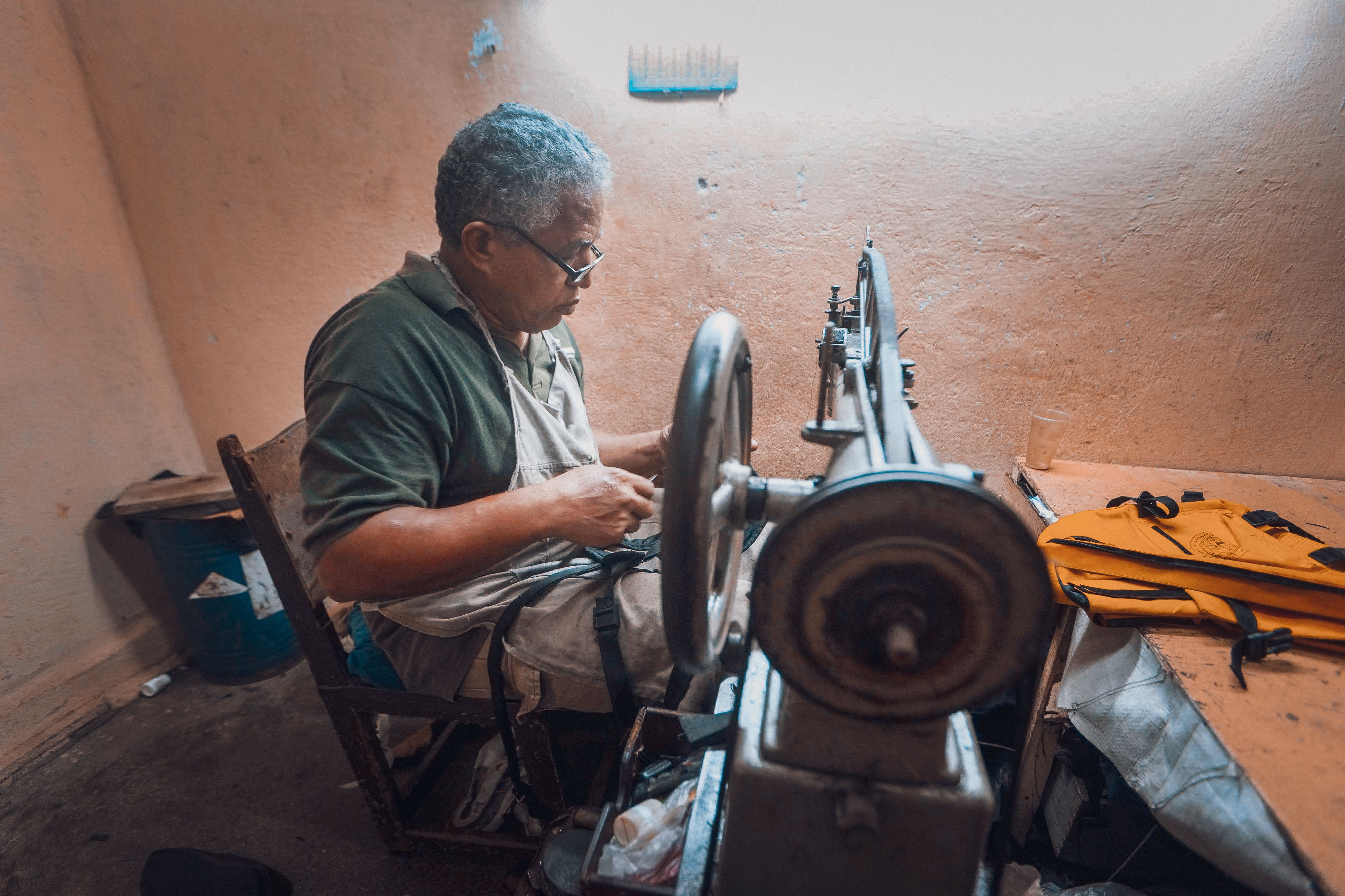 Elderly man sewing indoors using a vintage machine. Side view emphasizes his focused work.