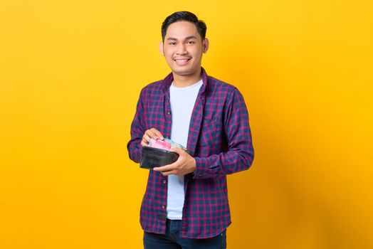 Happy young man in plaid shirt holding wallet with Indonesian rupiah against yellow background.