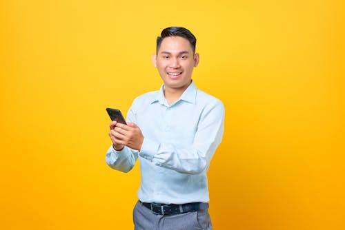 Man in Light Blue Long Sleeves Holding His Cellphone on Yellow Background