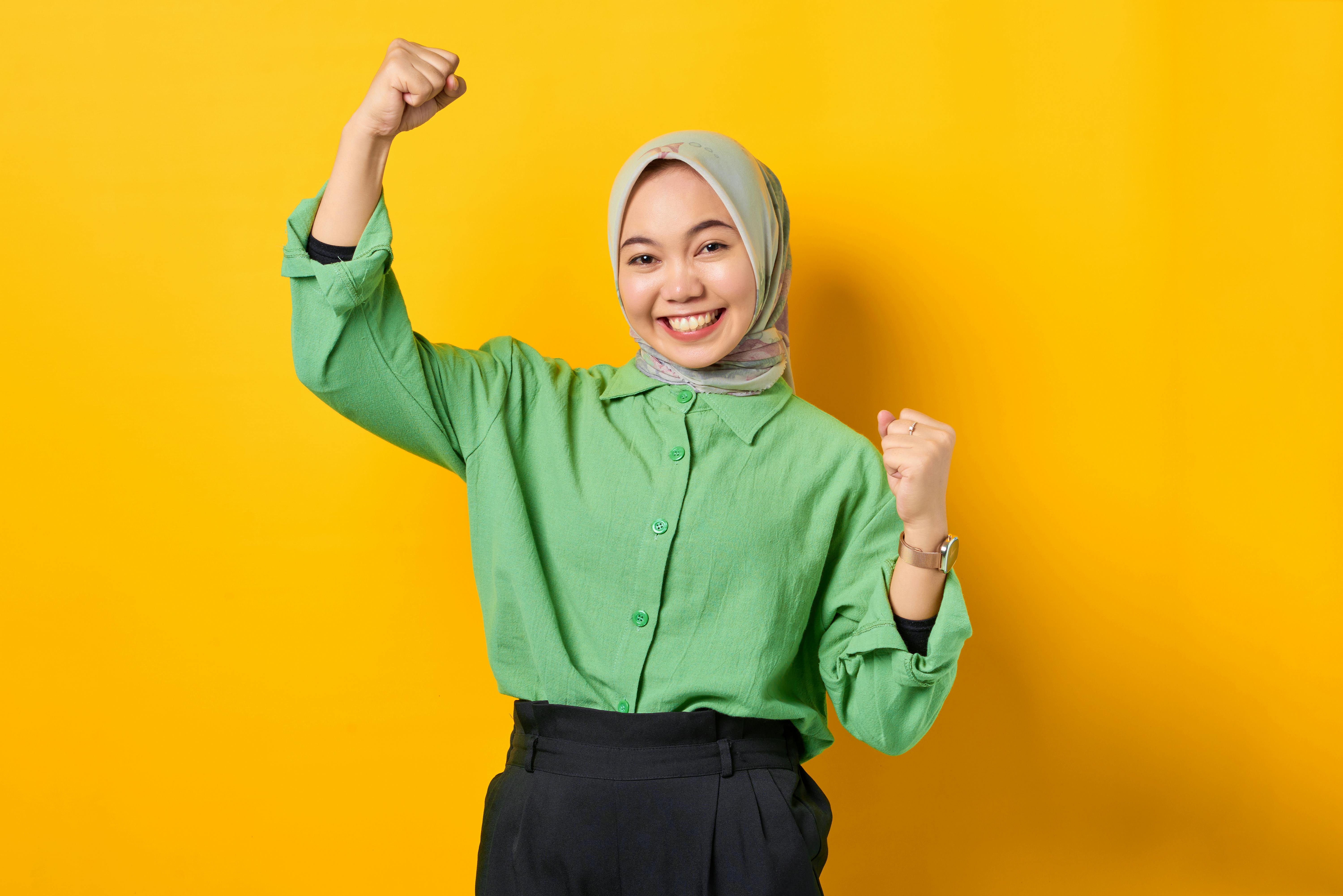 ahli SEO Indonesia - Joyful woman wearing a hijab and green shirt, smiling confidently against a bright yellow background.