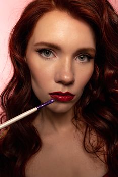 Close-up portrait of a beautiful redhead woman applying red lipstick, highlighting her flawless makeup and features.