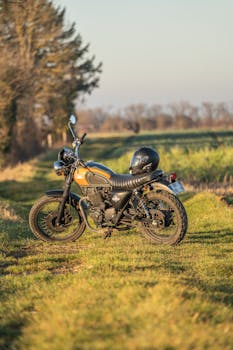 A classic motorcycle with helmet, parked on a rural grass path under a sunny sky.
