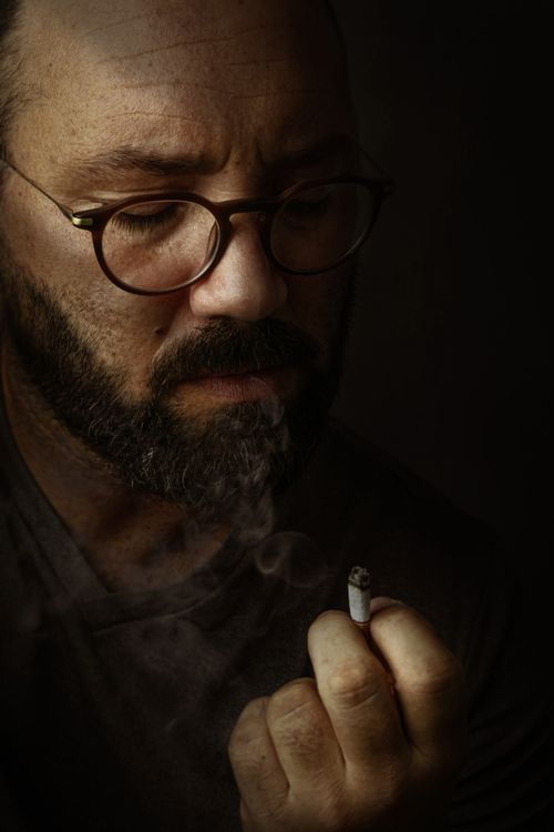Free Moody portrait of a bearded man with glasses, holding a cigarette in deep thought. Stock Photo