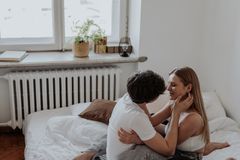 A loving couple sits and embraces on a bed in a cozy, sunlit bedroom.