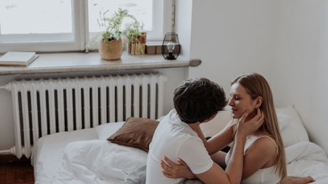 A loving couple sits and embraces on a bed in a cozy, sunlit bedroom.