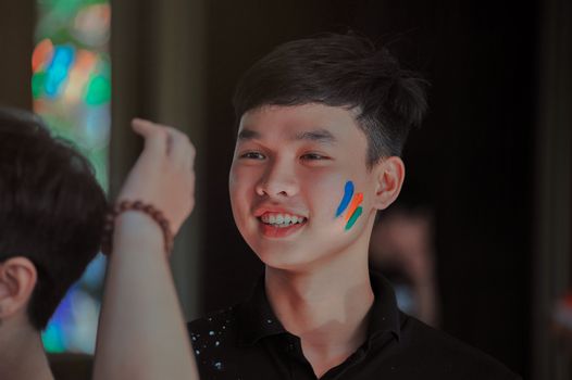 Young man with colorful face paint smiling indoors during a fun event.