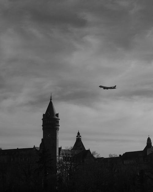 Grayscale Photo of Airplane Flying under Gloomy Sky