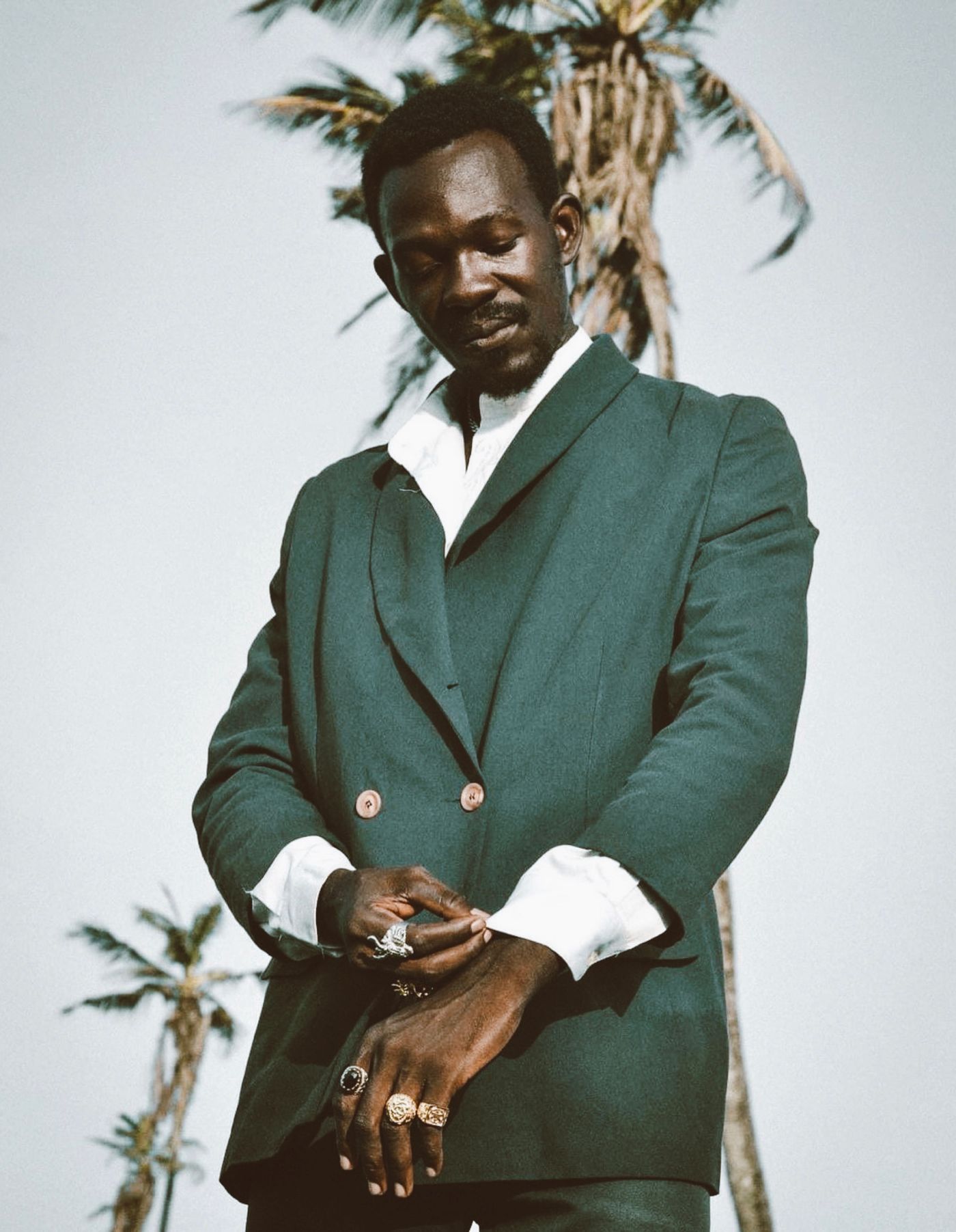 A man in a dark green suit adjusts his cuff beneath palm trees in Accra.