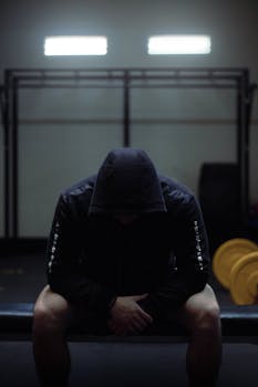 A man sitting on a bench in a dimly lit gym wearing a black hoodie, exuding mystery.