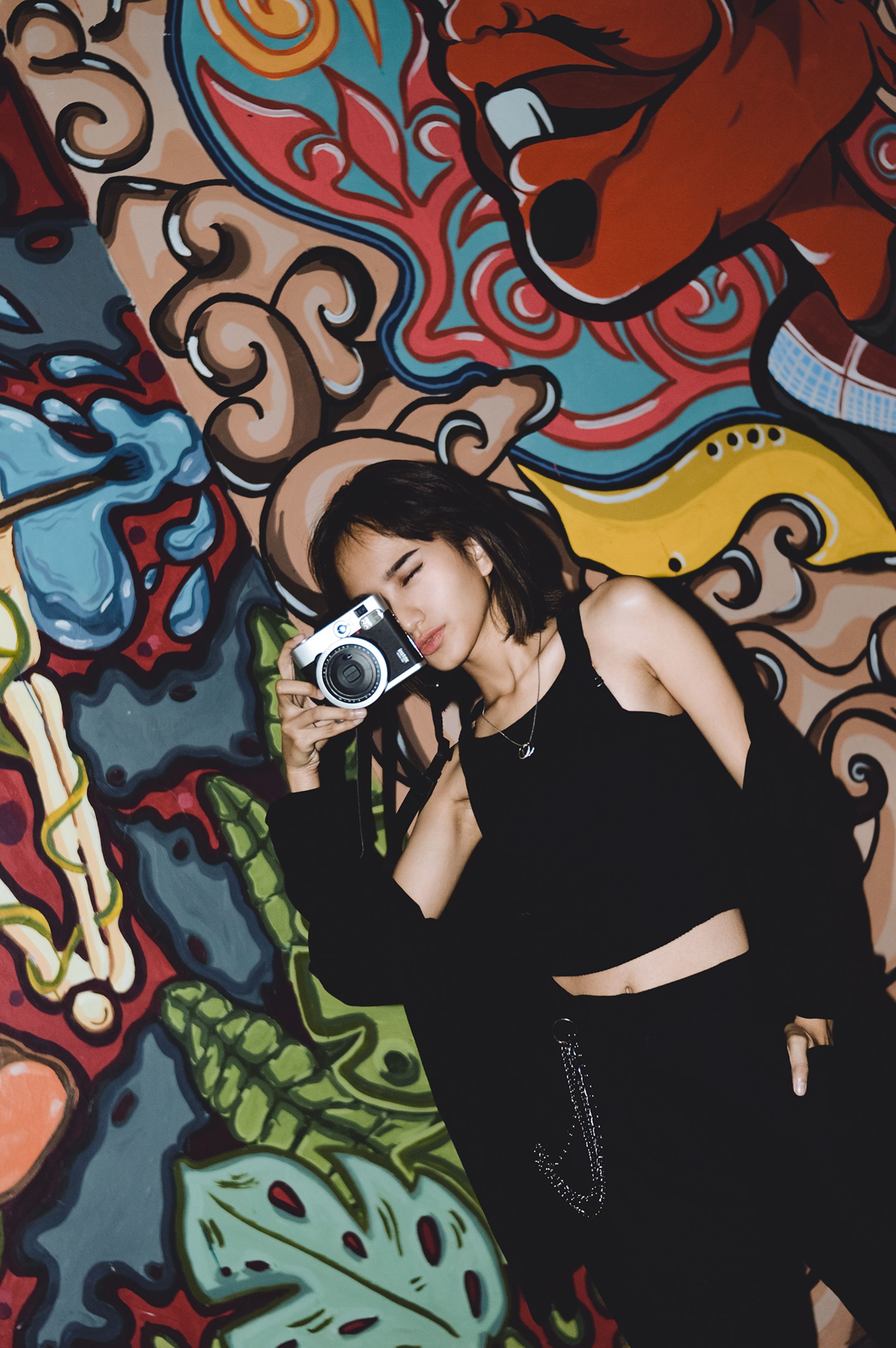 Stylish woman in black outfit poses with a camera against a vibrant graffiti wall, showcasing urban art.