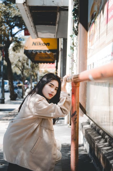 Asian woman posing on a sunny city street, leaning gracefully on a wall.