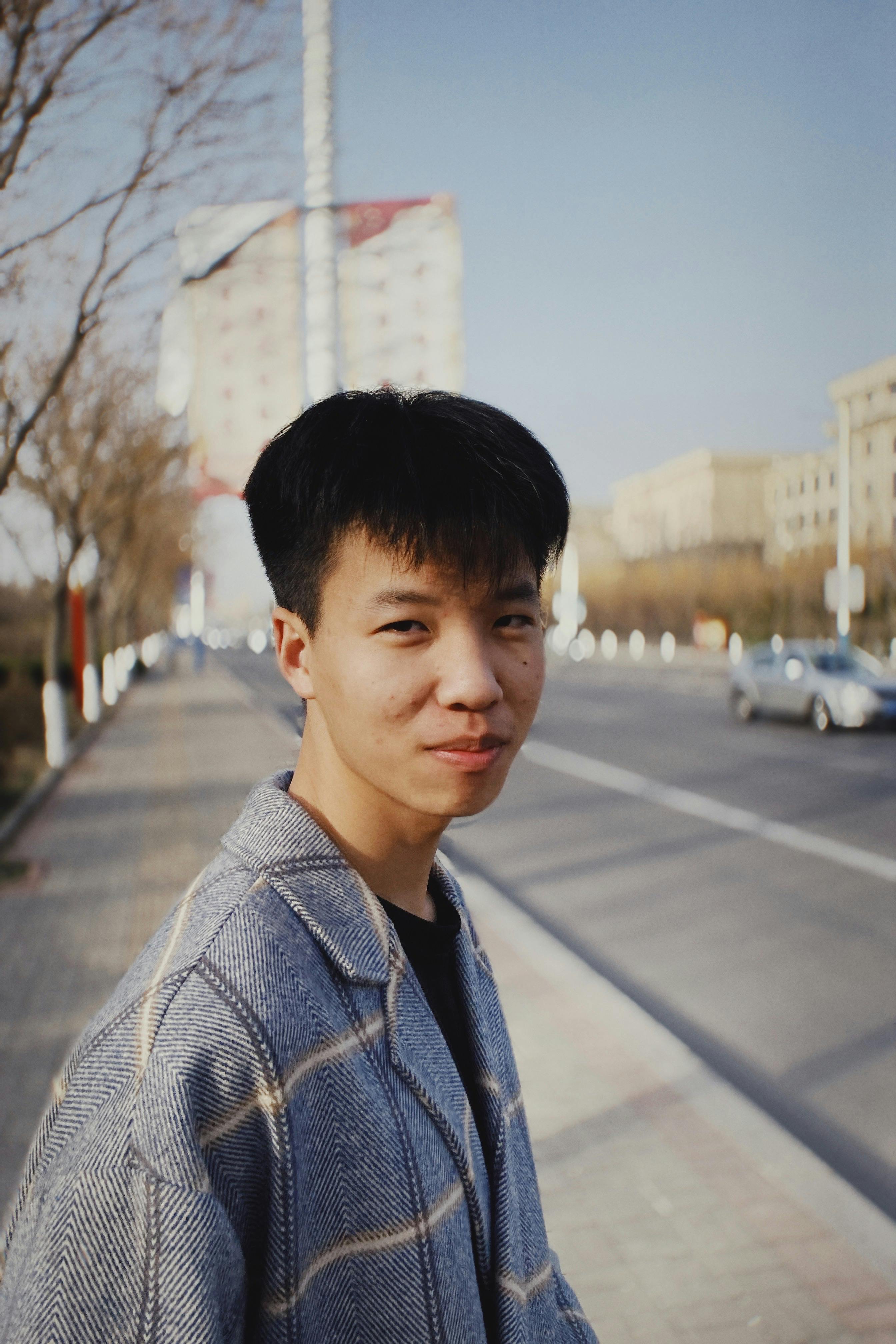 A young man in a checkered coat stands on a city street, smiling naturally.