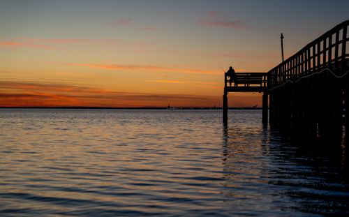 Free Silhouette of a person fishing on a pier during a stunning orange sunset over calm waters. Stock Photo