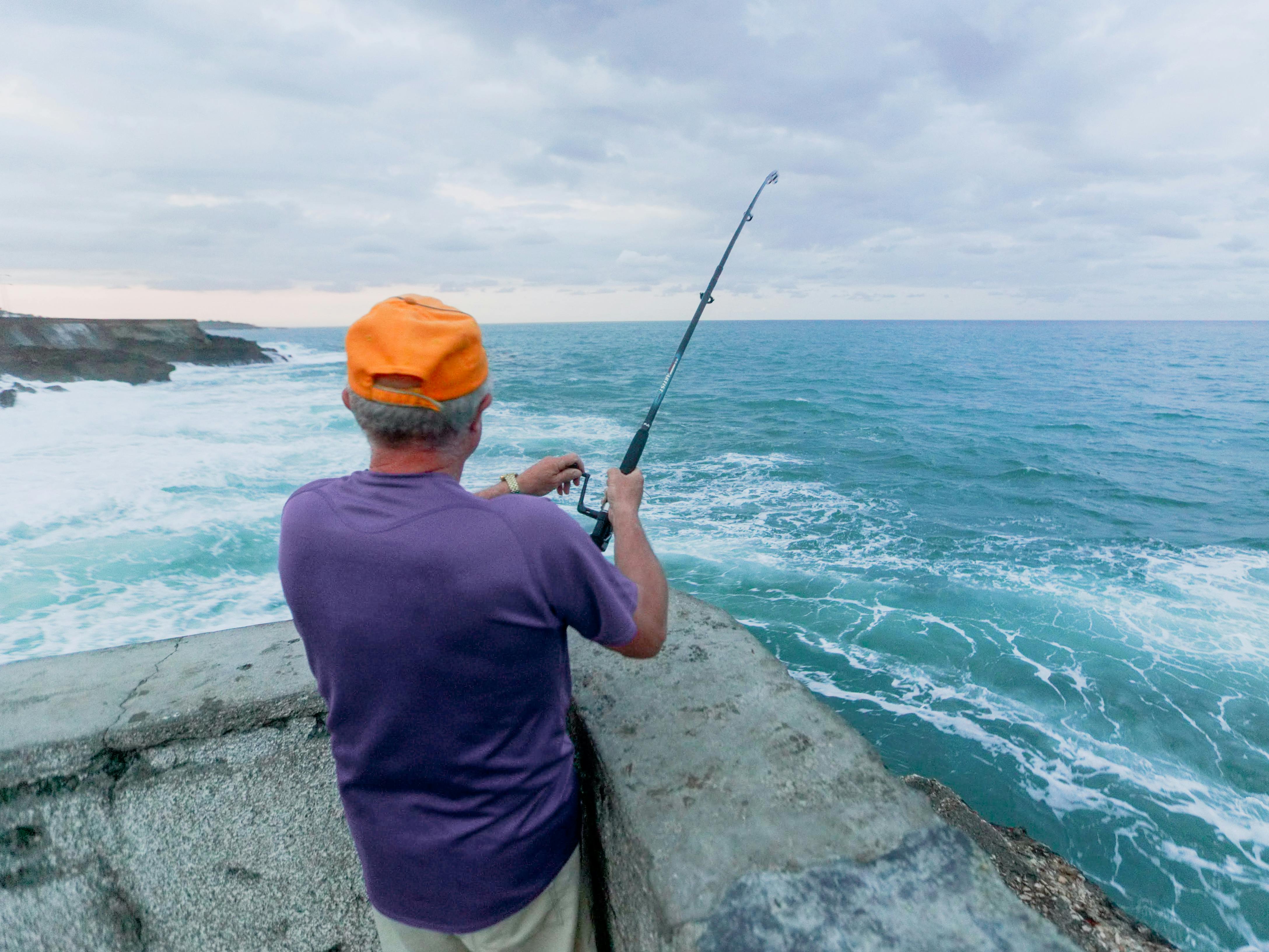 Man in Purple Shirt and Orange Cap Holding Fishing Rod Near Sea