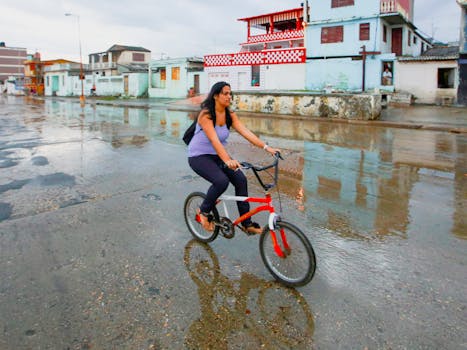 A woman rides a bicycle on a wet street in an urban setting after rainfall, reflecting colorful buildings.
