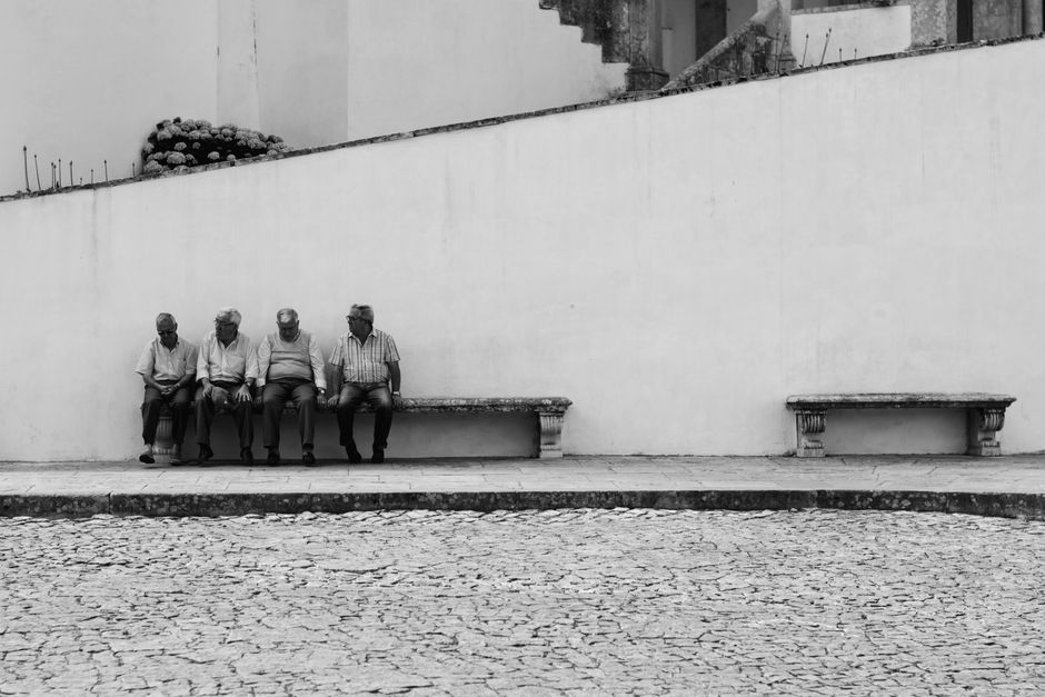 Four elderly men sitting on benches by a stone wall, outdoors in a quiet setting.