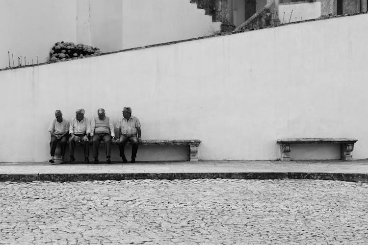 Grayscale Photo Of Four Men Sitting On Bench Along The Street